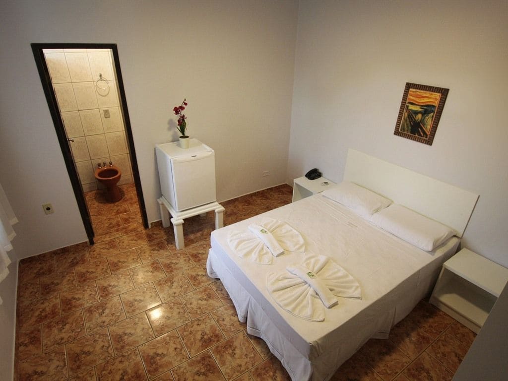 Une chambre d’hôtel avec un lit, une table de chevet, une petite table avec un vase, des toilettes et un tableau au mur.
