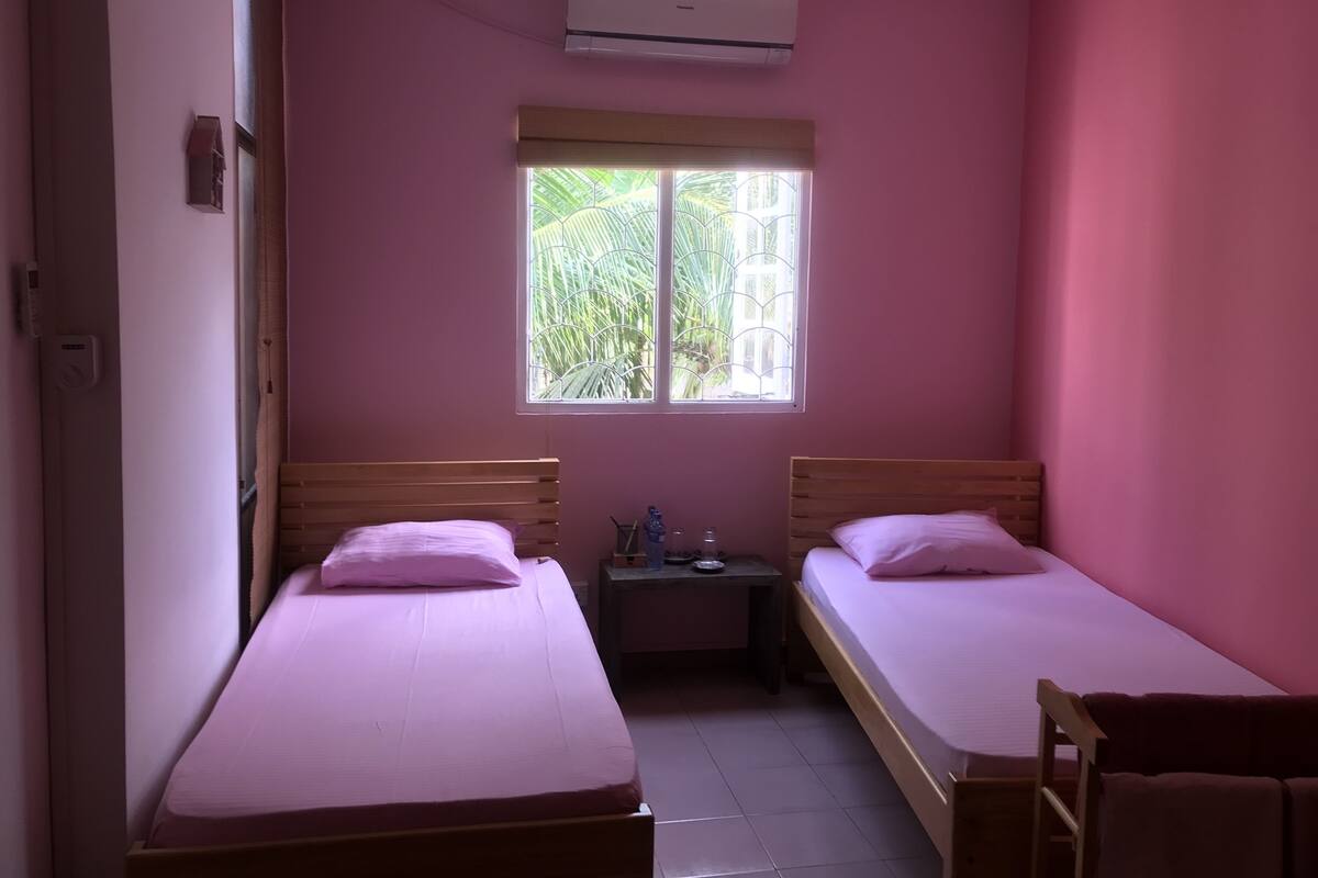 Une petite chambre à coucher décorée dans des tons roses, avec deux lits simples, une table de chevet et une fenêtre donnant sur un paysage verdoyant.
