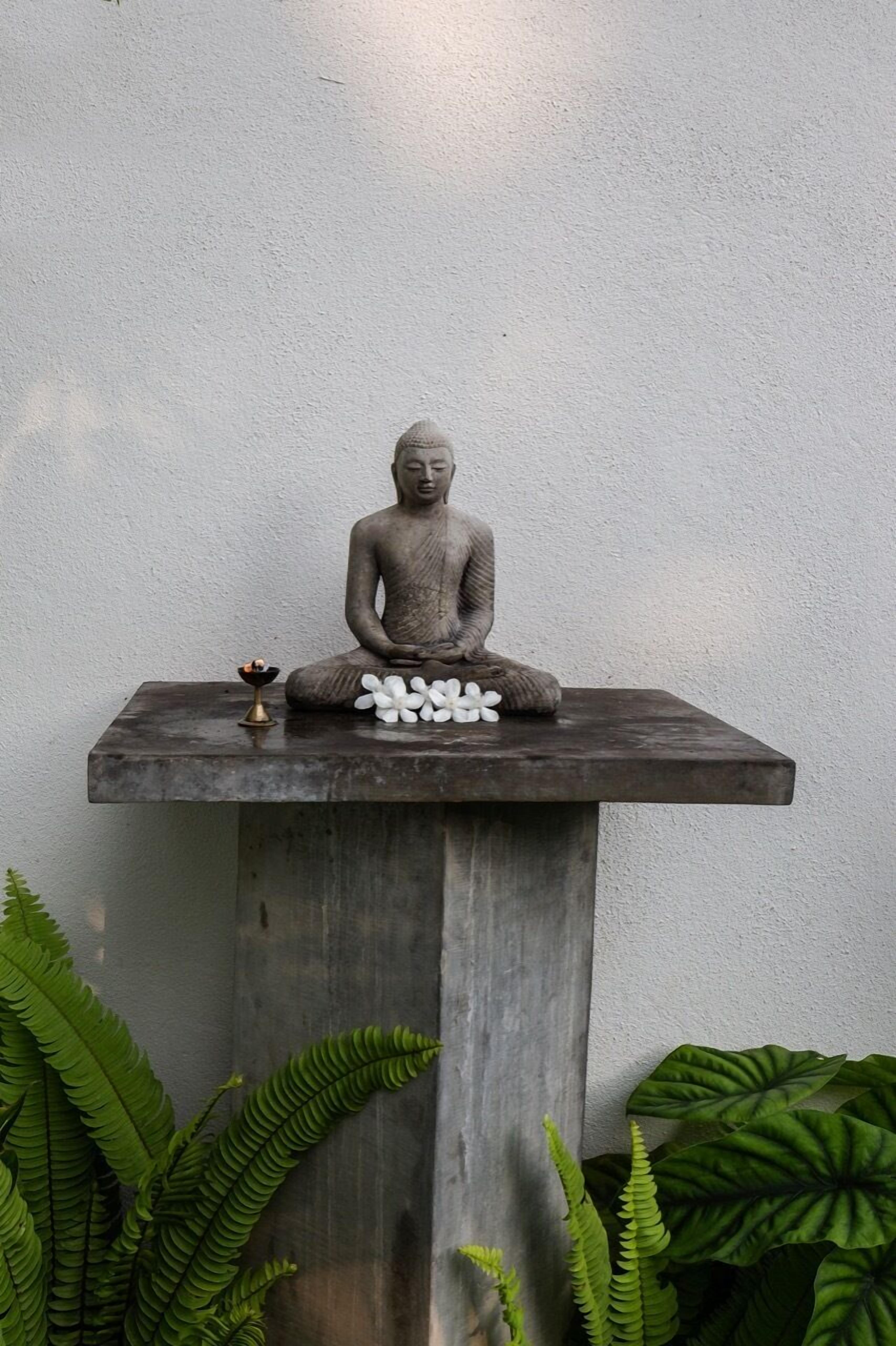 A stone Buddha statue on a wooden platform with white flowers and a small incense burner.