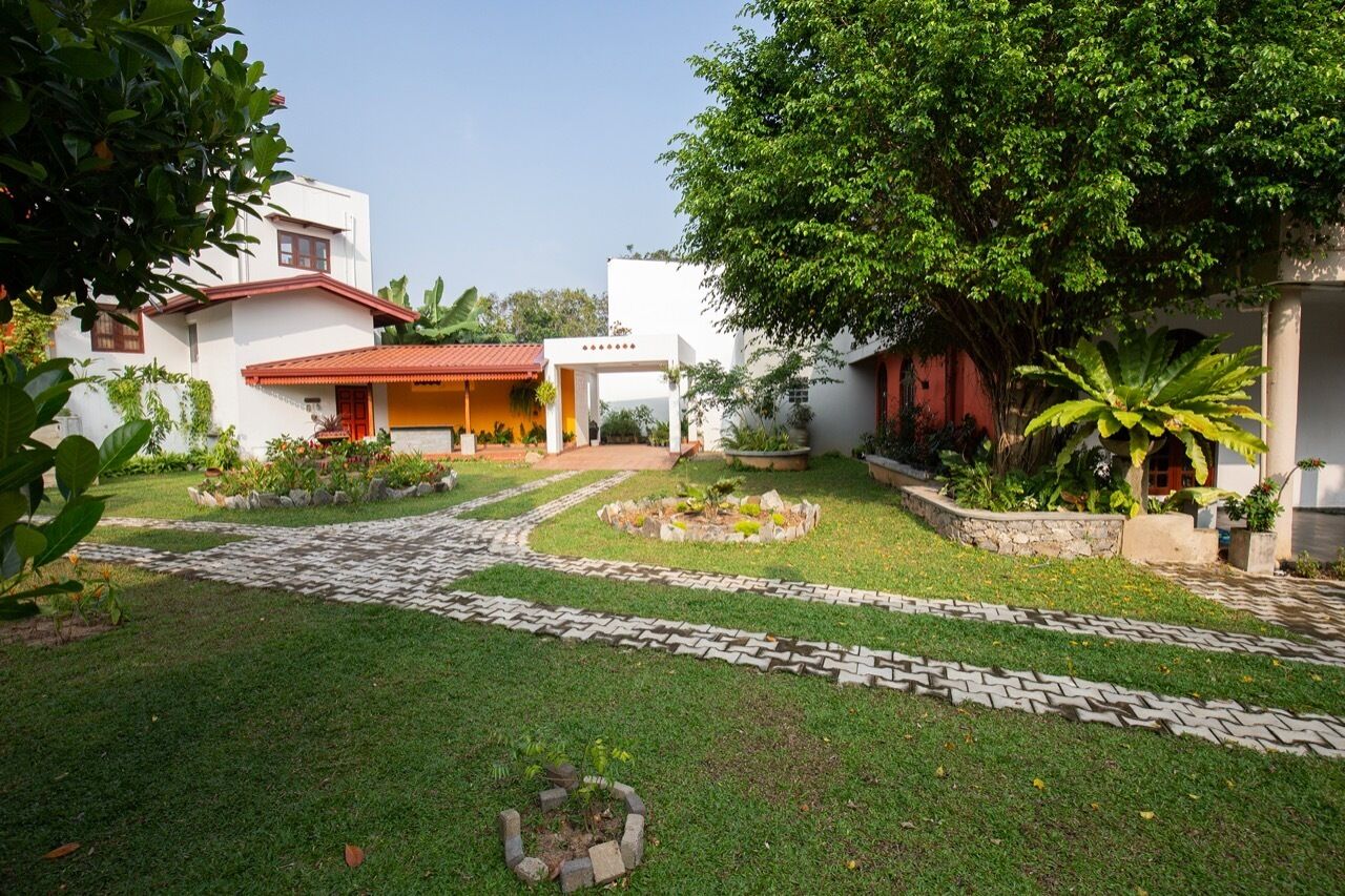 Un patio con un camino pavimentado, una zona de estar techada y diversas plantas y árboles.