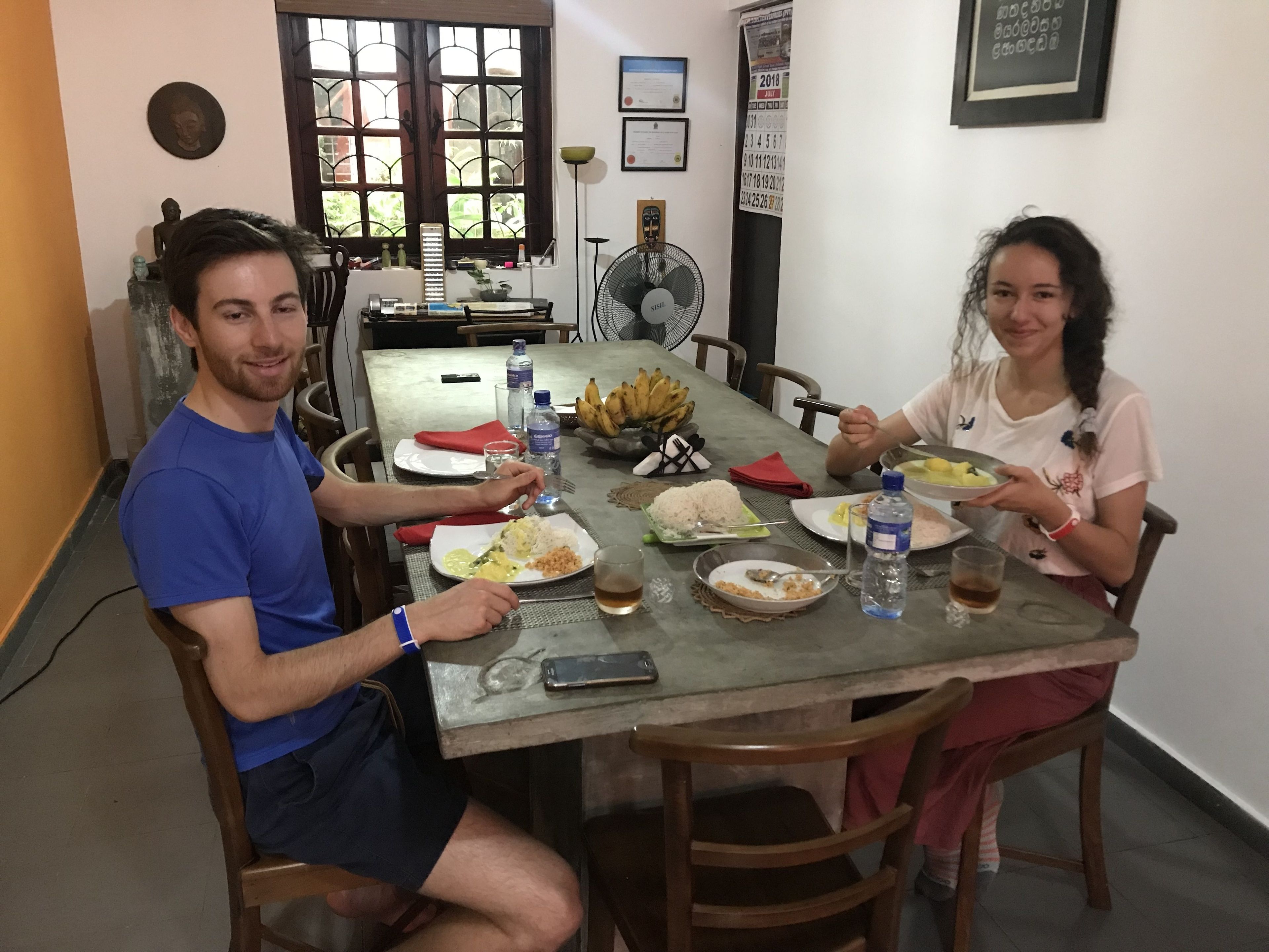 A dining area with a table set for two, featuring plates of food, a fan, and a calendar on the wall.