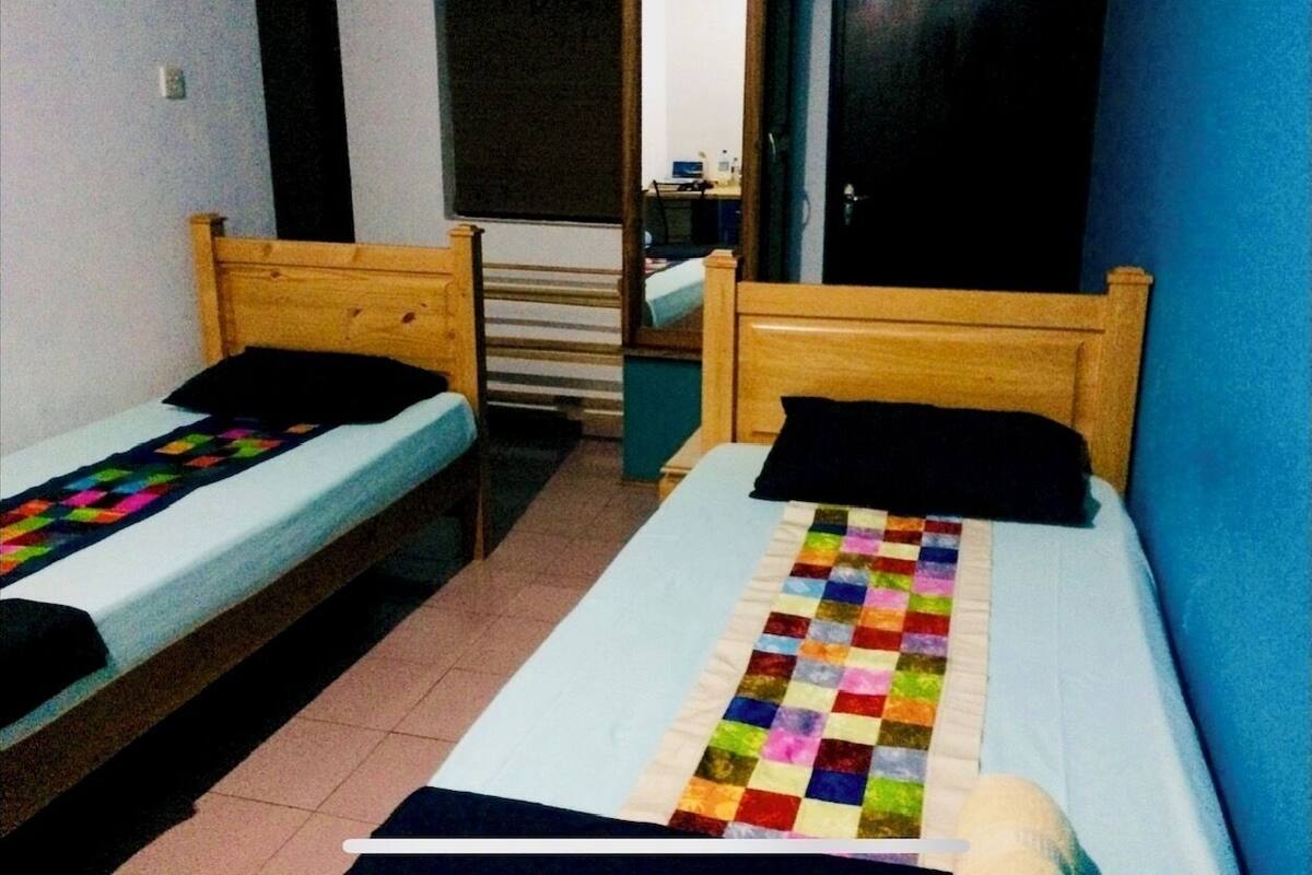 Une petite chambre avec deux lits simples, une tête de lit en bois, une courtepointe aux couleurs vives et un miroir au mur.