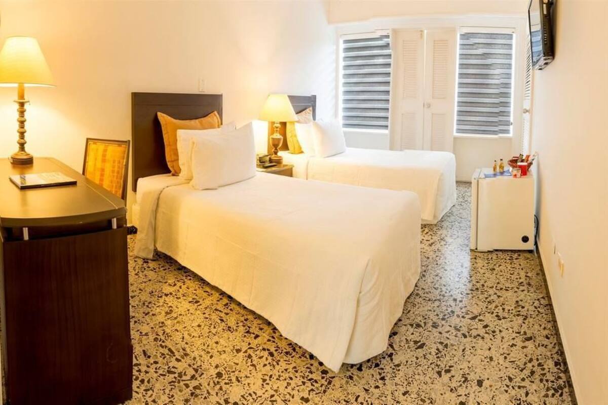 Ein Hotelzimmer mit zwei Betten, einem kleinen Nachttisch mit Lampe, einem an der Wand befestigten Fernseher und einem Fenster mit Jalousien.