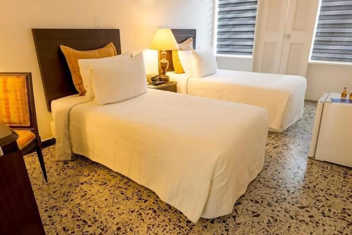 Ein Hotelzimmer mit zwei Betten, einem Nachttisch mit Lampe, einem gerahmten Bild und einem Fenster mit Jalousien.