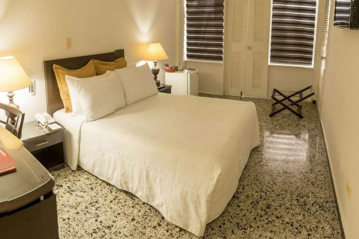 Ein Hotelzimmer mit einem Bett, zwei Nachttischen mit Lampen, einem Sessel, einem kleinen Tisch mit Buch und Telefon sowie einem Fenster mit Jalousien.