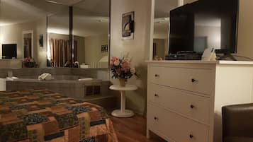 A hotel room with a large bed, a sitting area with a chair, a desk with a computer, a large mirror, a TV, and a small table with a vase of flowers.