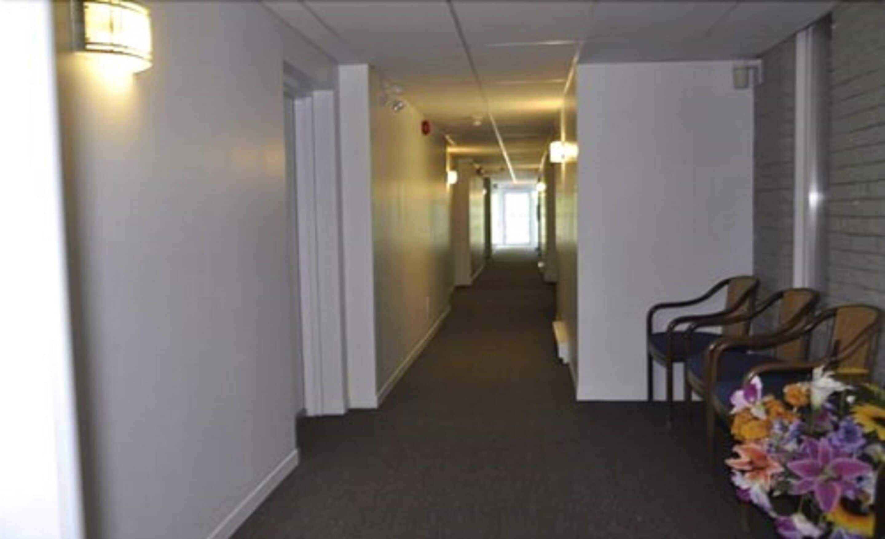 A hotel hallway with a carpeted floor, white walls, and a row of wooden chairs on the right.