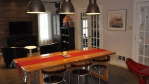 Un comedor moderno con una mesa de madera, taburetes de bar, iluminación colgante y un televisor al fondo.