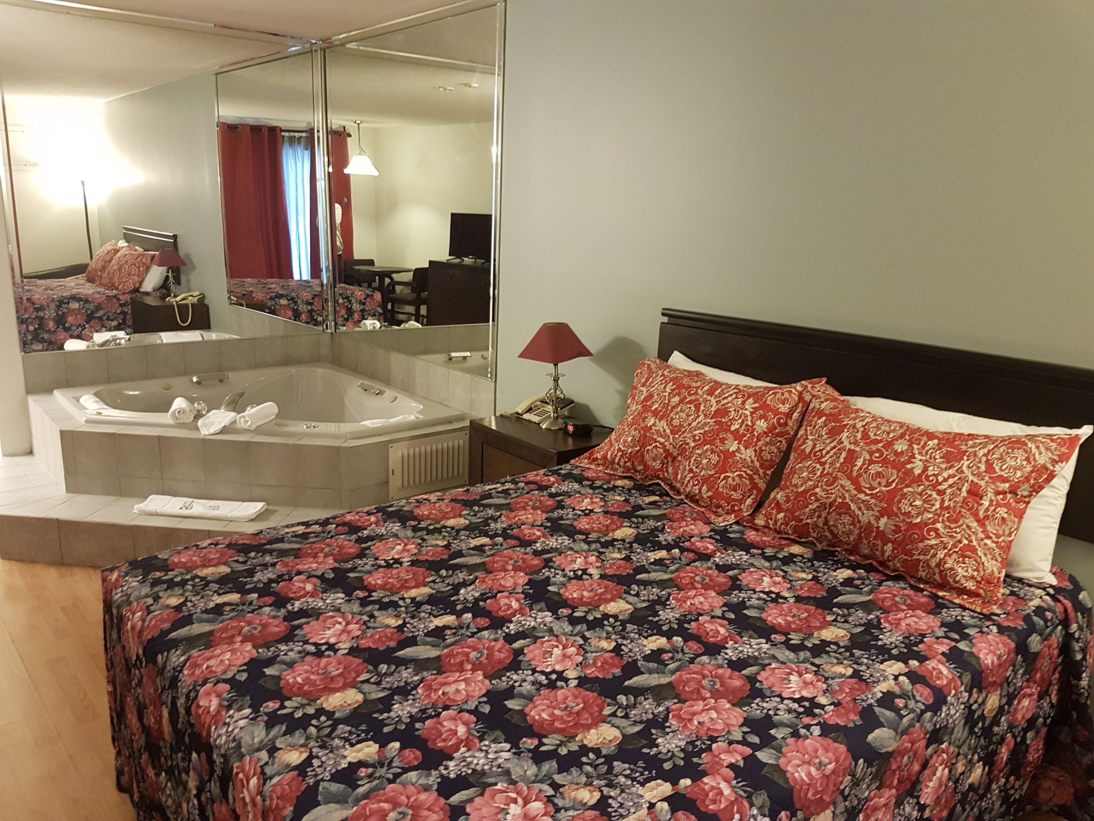 A hotel room with a large bed, a sitting area with a chair and a table, a large mirror, a bathtub, and a television.