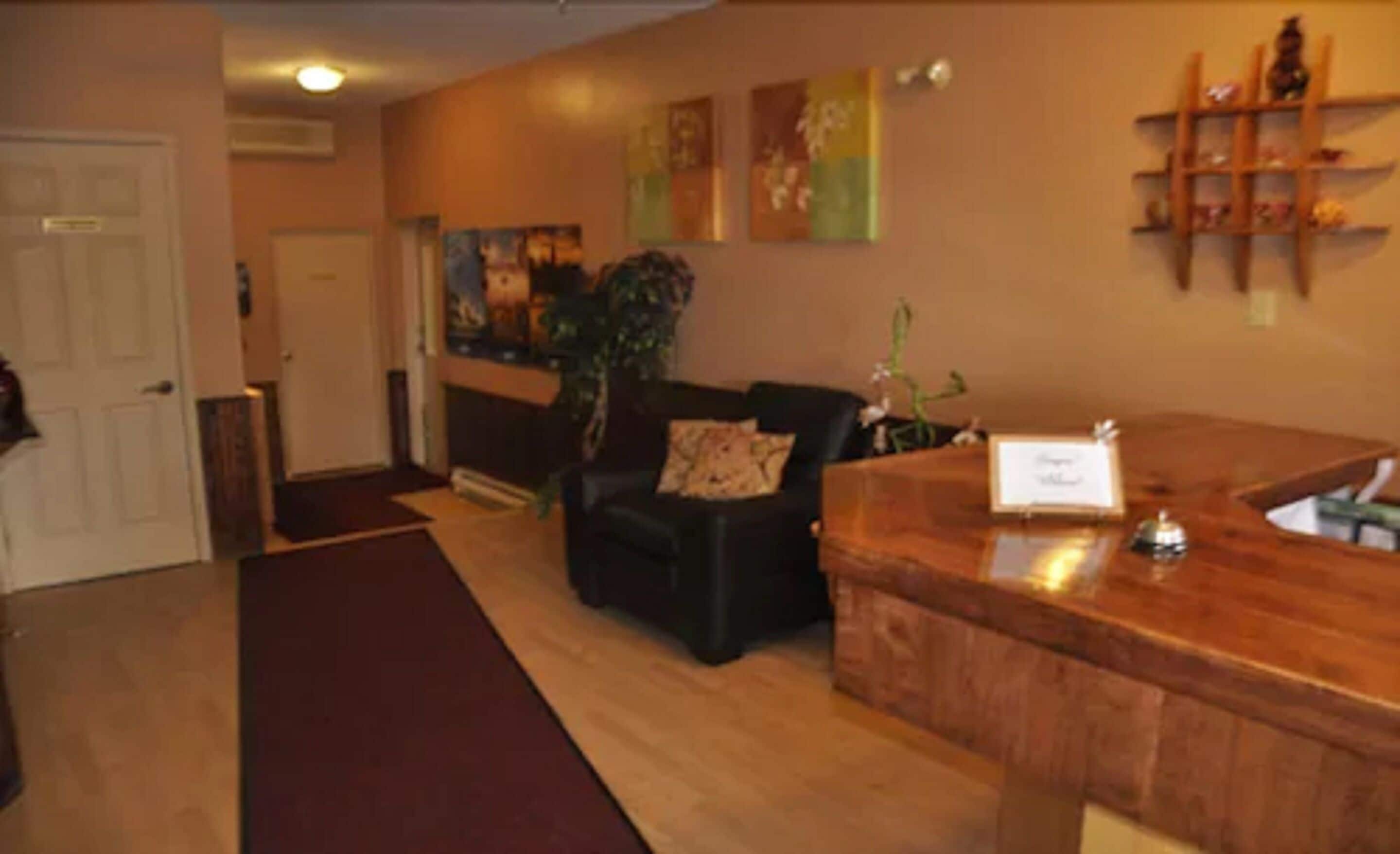 Un espace d’accueil avec un comptoir de réception en bois, un canapé en cuir noir agrémenté d’un coussin, une plante et une porte en arrière-plan.