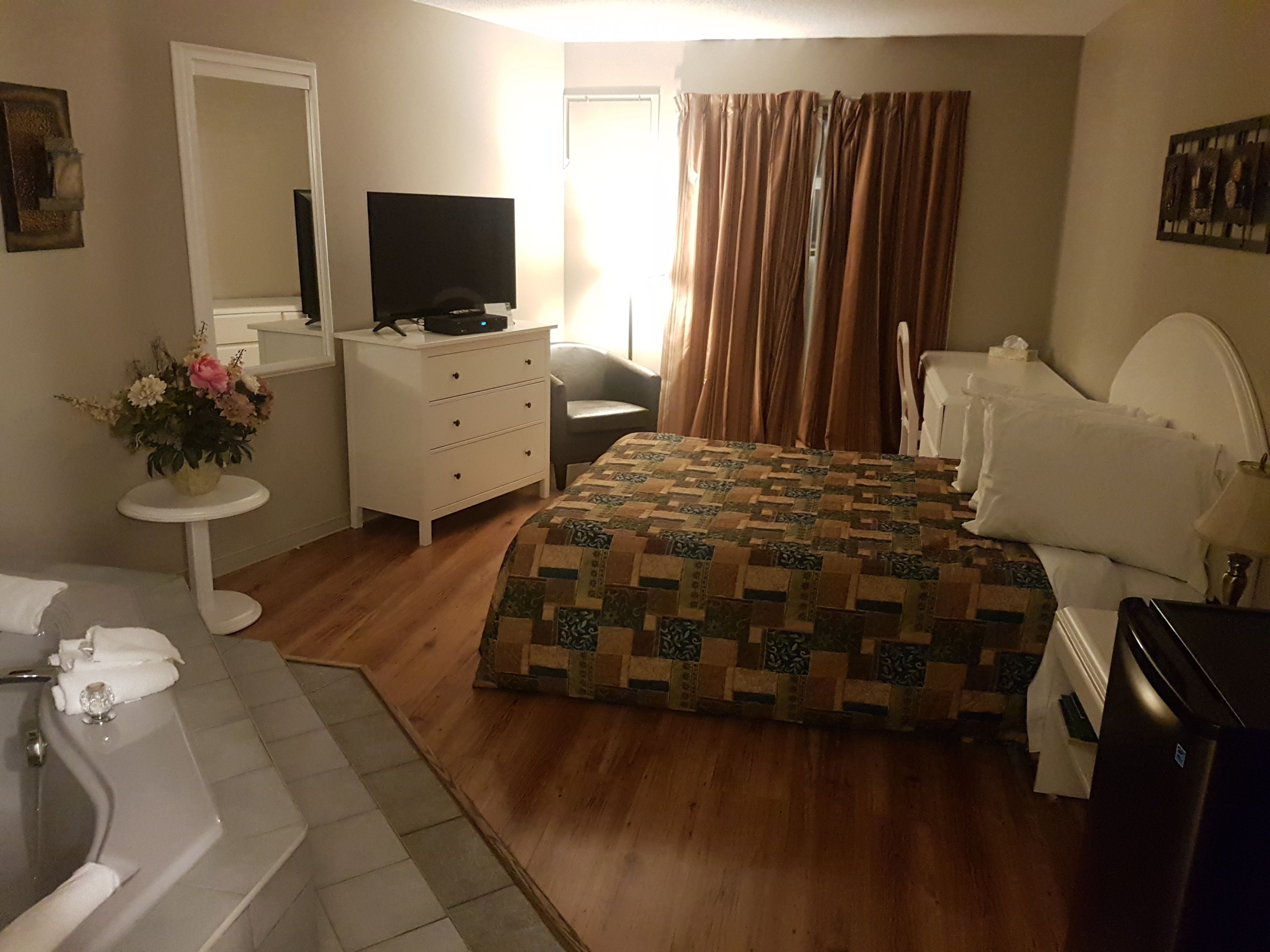 A bedroom with a bed, a dresser, a television, a bathroom with a bathtub, and a window with curtains.