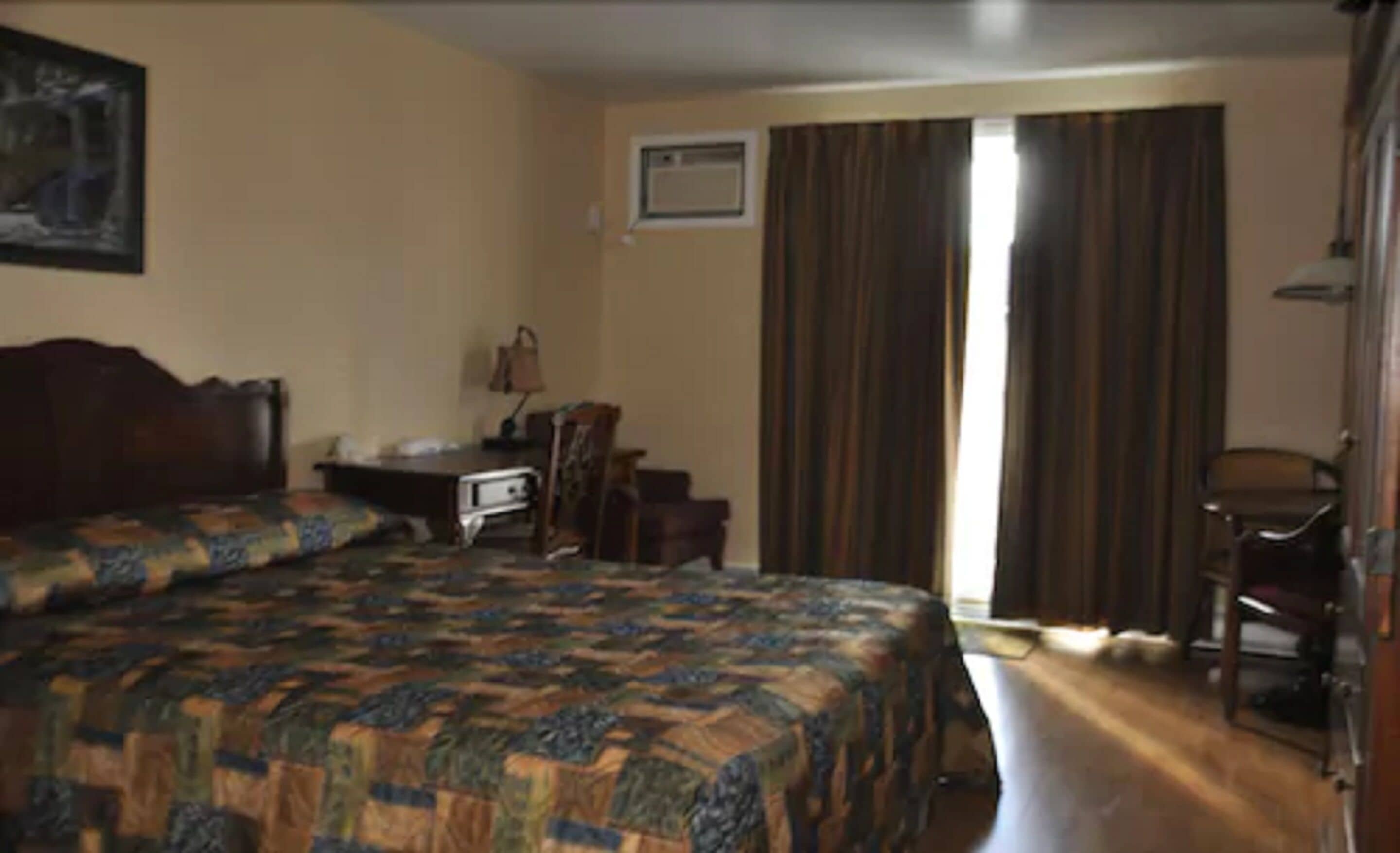 A hotel room with a large bed, a desk with a chair, a window with curtains, and a door.