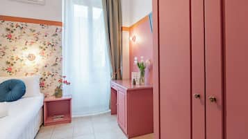 Ein Zimmer mit floraler Tapete, einem weißen Sofa mit blauen Kissen, einem rosa Schreibtisch mit Blumenvase und einem rosa Kleiderschrank.