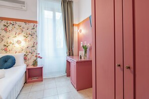 A room with a floral wallpaper, a white sofa with blue cushions, a pink desk with a vase of flowers, and a pink wardrobe. - España (Rome)