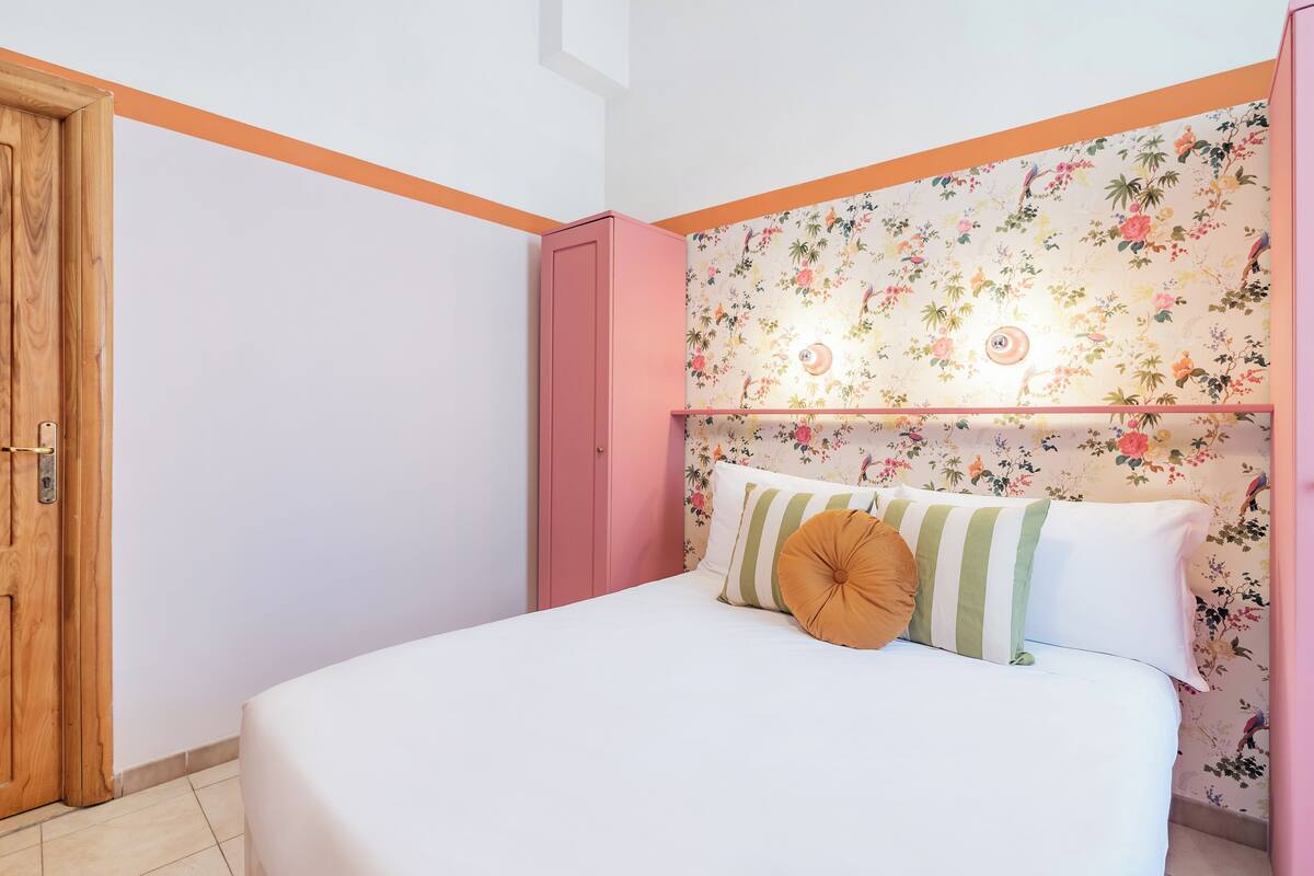 A bedroom with a floral wallpaper headboard, a bed with white bedding, and a pink wardrobe.