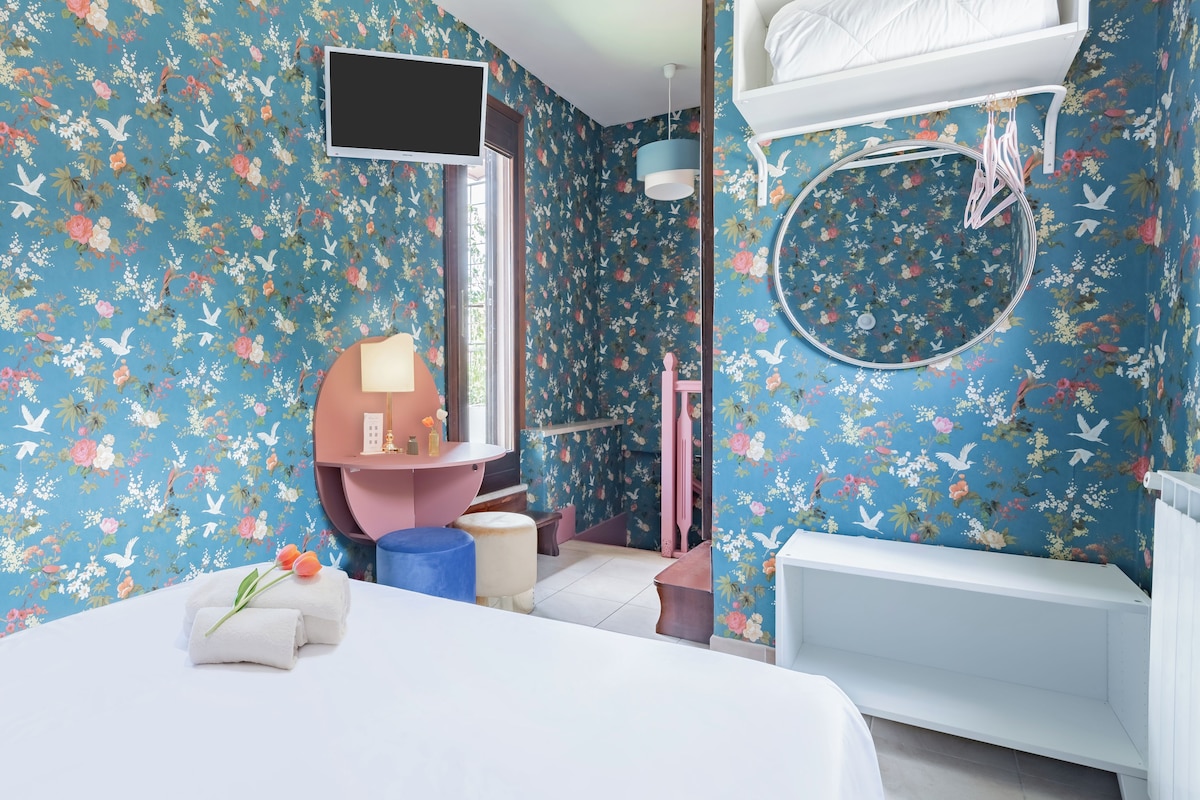 A bedroom with floral wallpaper, a bed, a bedside table, a mirror, a hanging light, a window, and a bathroom with a bathtub and toilet.
