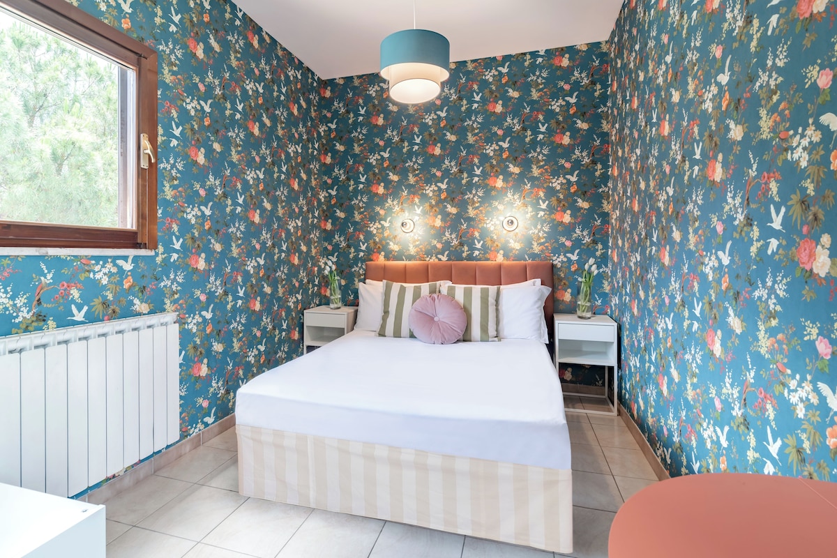 A bedroom with a floral wallpaper, a bed with a striped bedspread, two bedside tables with lamps, and a window with a view of trees.