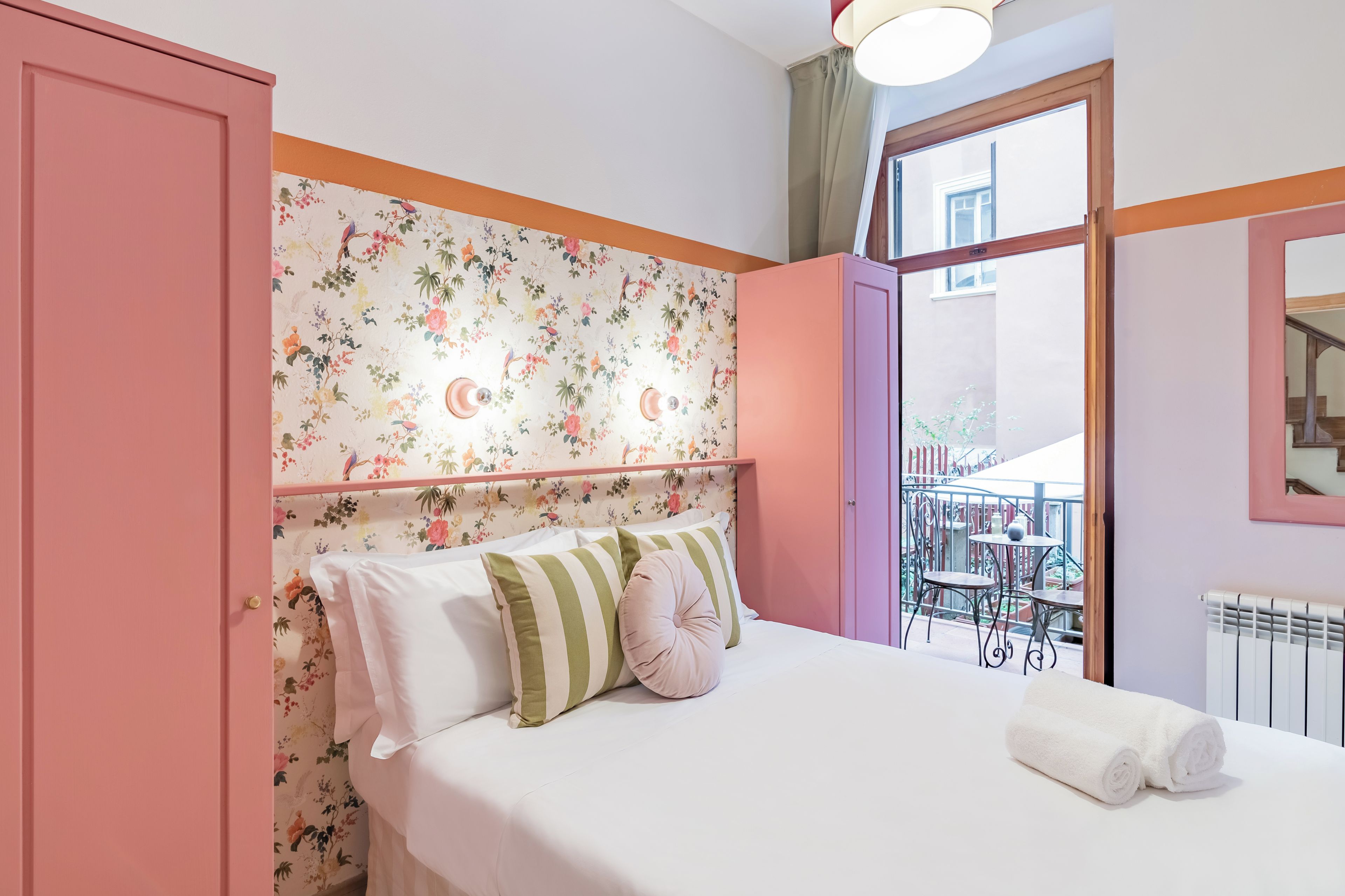 Ein Schlafzimmer mit floraler Tapete, einem Bett mit weißer Bettwäsche und gestreifter Bettwäsche, einer rosafarbenen Tür und einem Balkon mit Tisch und Stühlen.