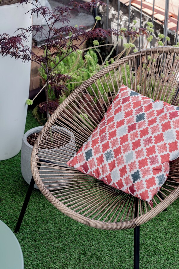 Una silla de mimbre con un cojín estampado sobre césped artificial, rodeada de plantas en macetas.