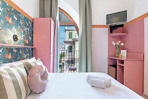A hotel room with a bed, a TV mounted on the wall, a small table with a chair, and a balcony with a view of buildings. - España (Rome)