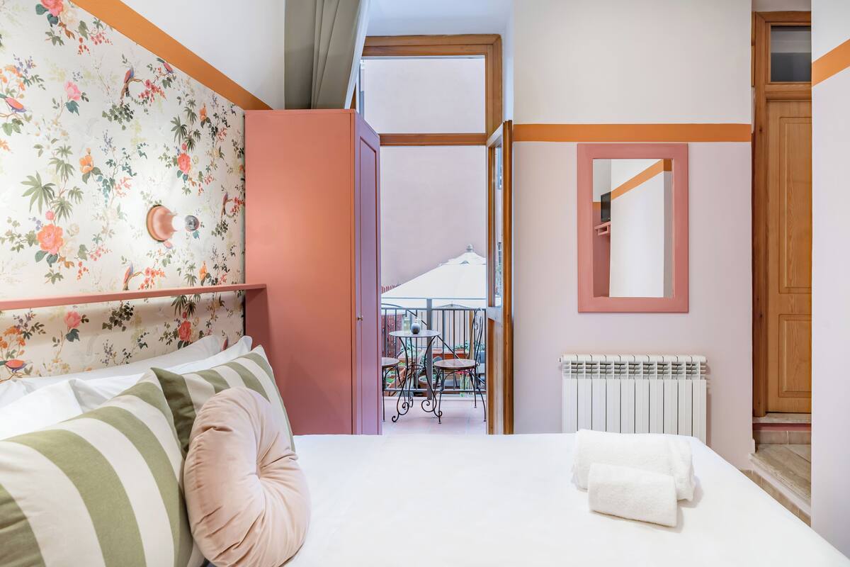 A bedroom with a bed, pillows, a radiator, a mirror, and a balcony with a table and chairs.