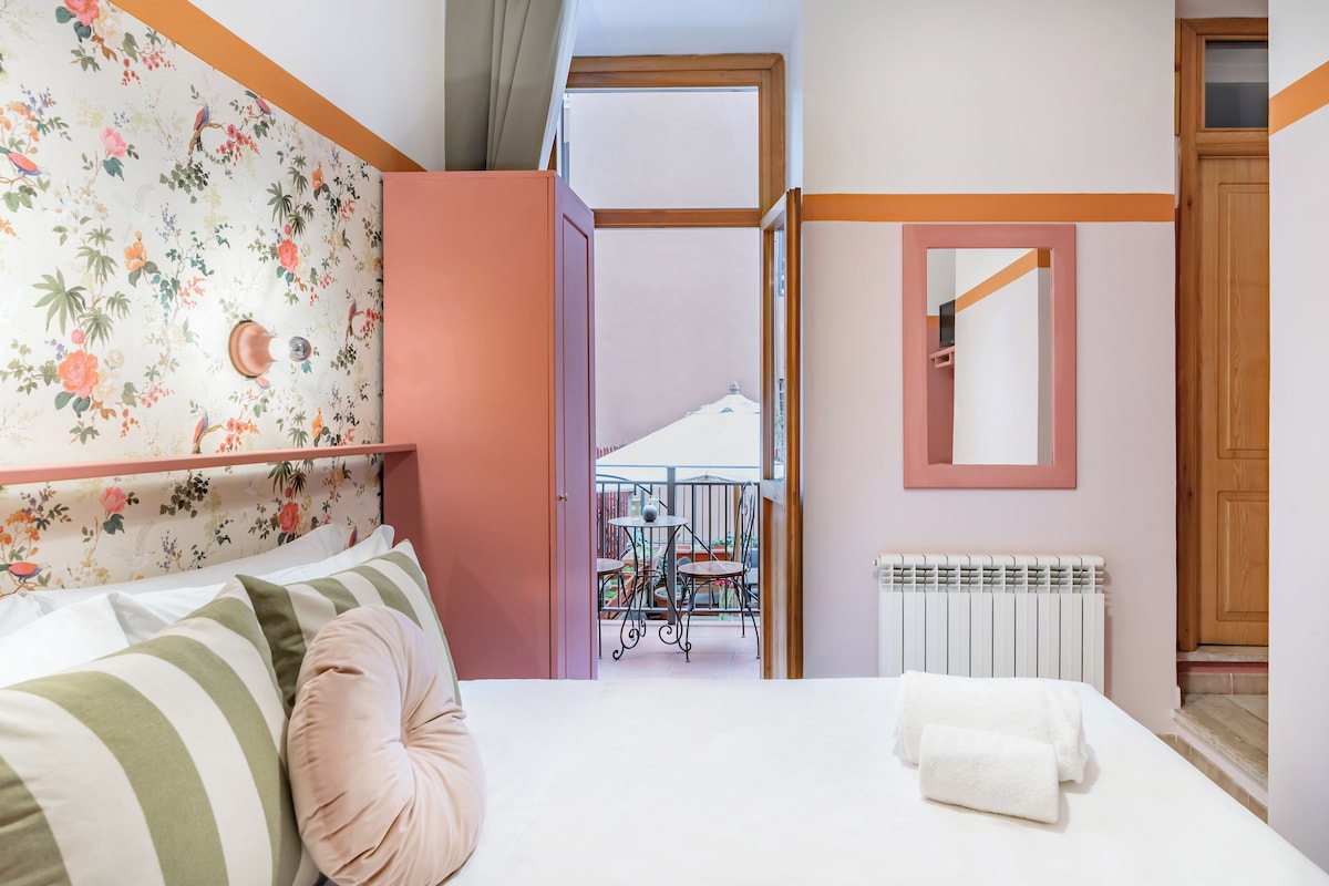 A bedroom with a bed, pillows, a radiator, a mirror, and a balcony with a table and chairs.