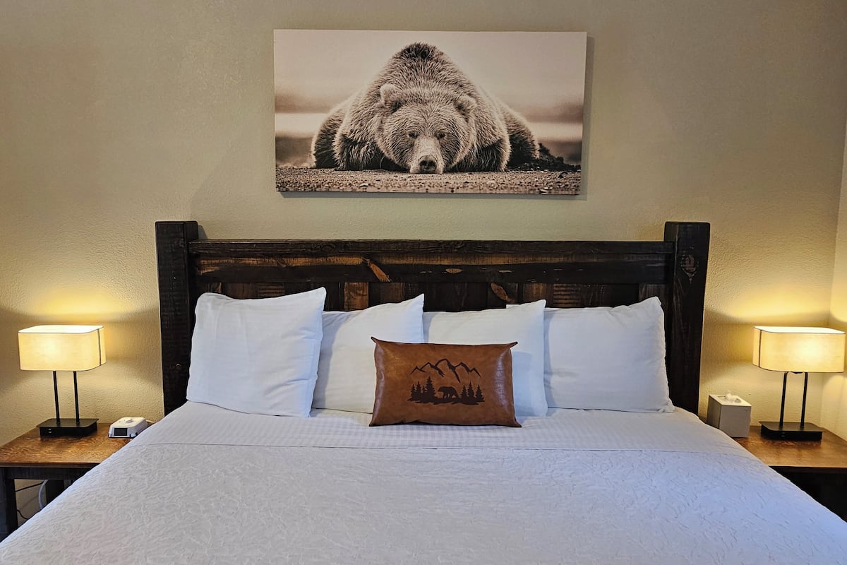 Una cama bien hecha con sábanas blancas y un cojín marrón con diseño de oso y árboles, flanqueada por dos lámparas de noche sobre mesitas de madera.