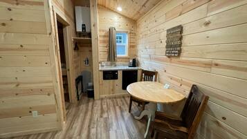 Interior de una cabaña de madera con una mesa de comedor y sillas, una zona de cocina con ventana, y piso de madera.