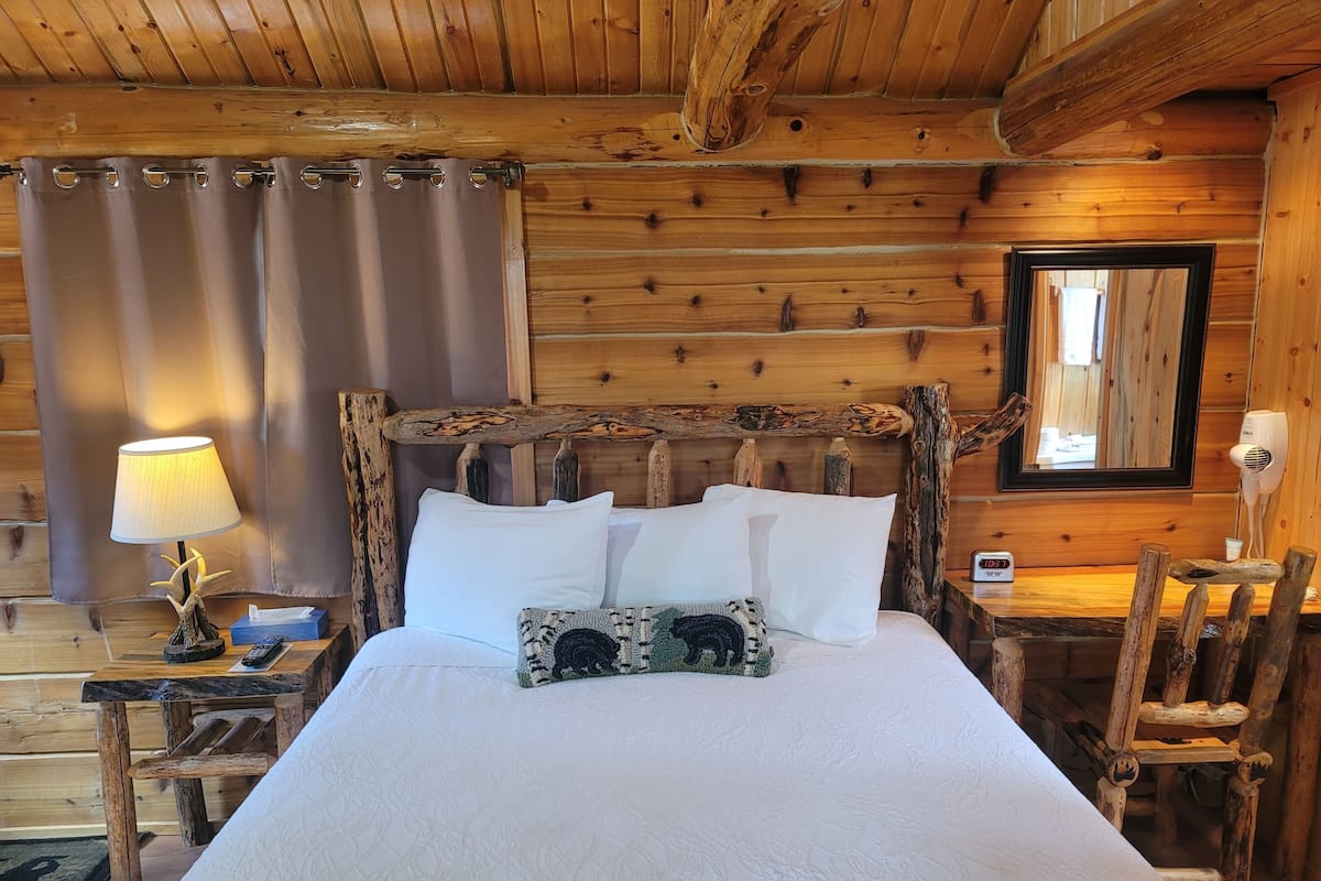 Un dormitorio de cabaña de madera con una cama, una mesita de noche con lámpara, un espejo y una silla.