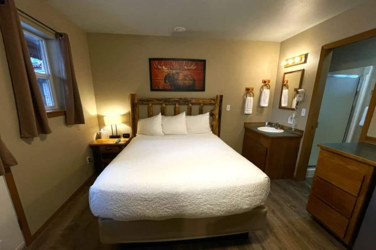 Un dormitorio con una cama grande, cabecera de madera, mesita de noche con lámpara, una mesita más pequeña con un teléfono, un espejo y cómoda de madera.