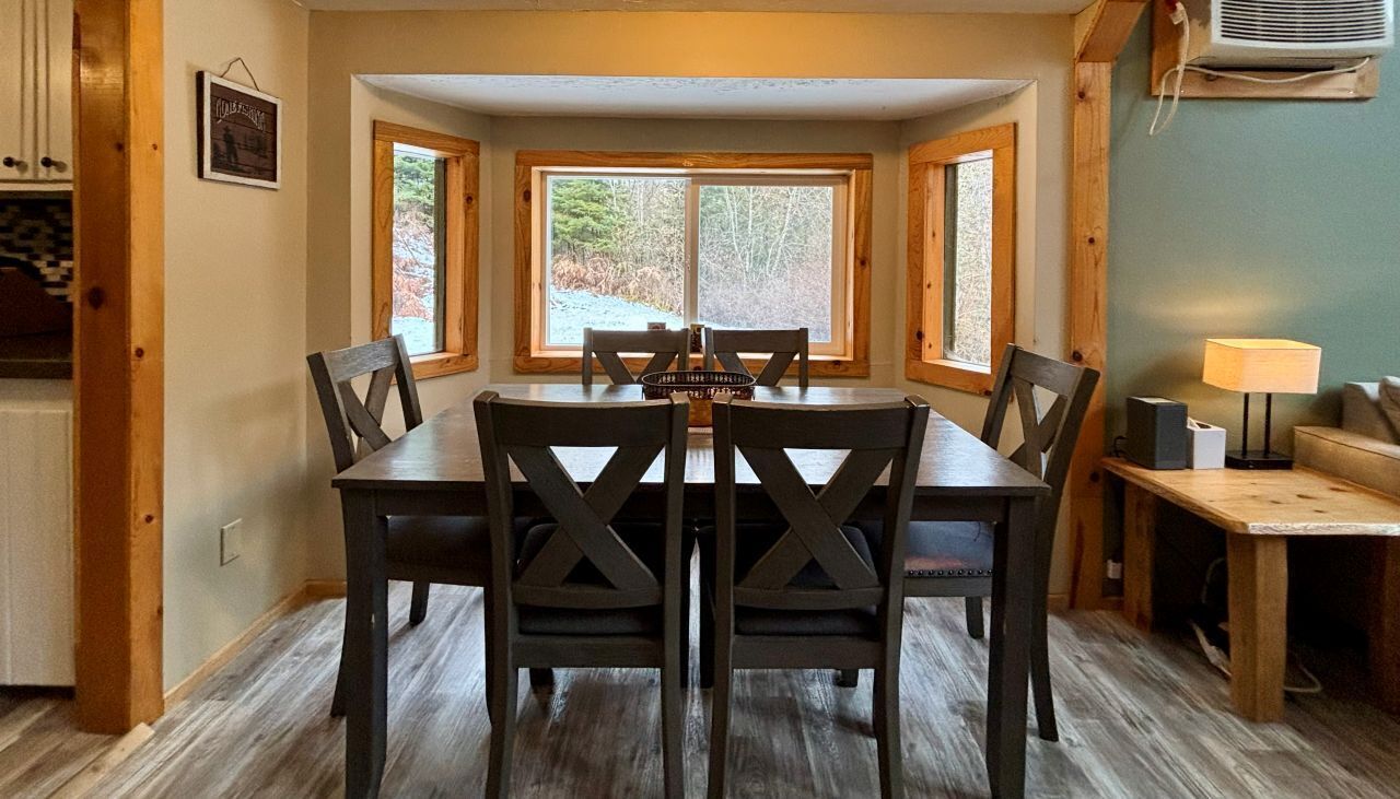 A dining area with a wooden table and chairs, a window with a view of trees, and a lamp on a side table.