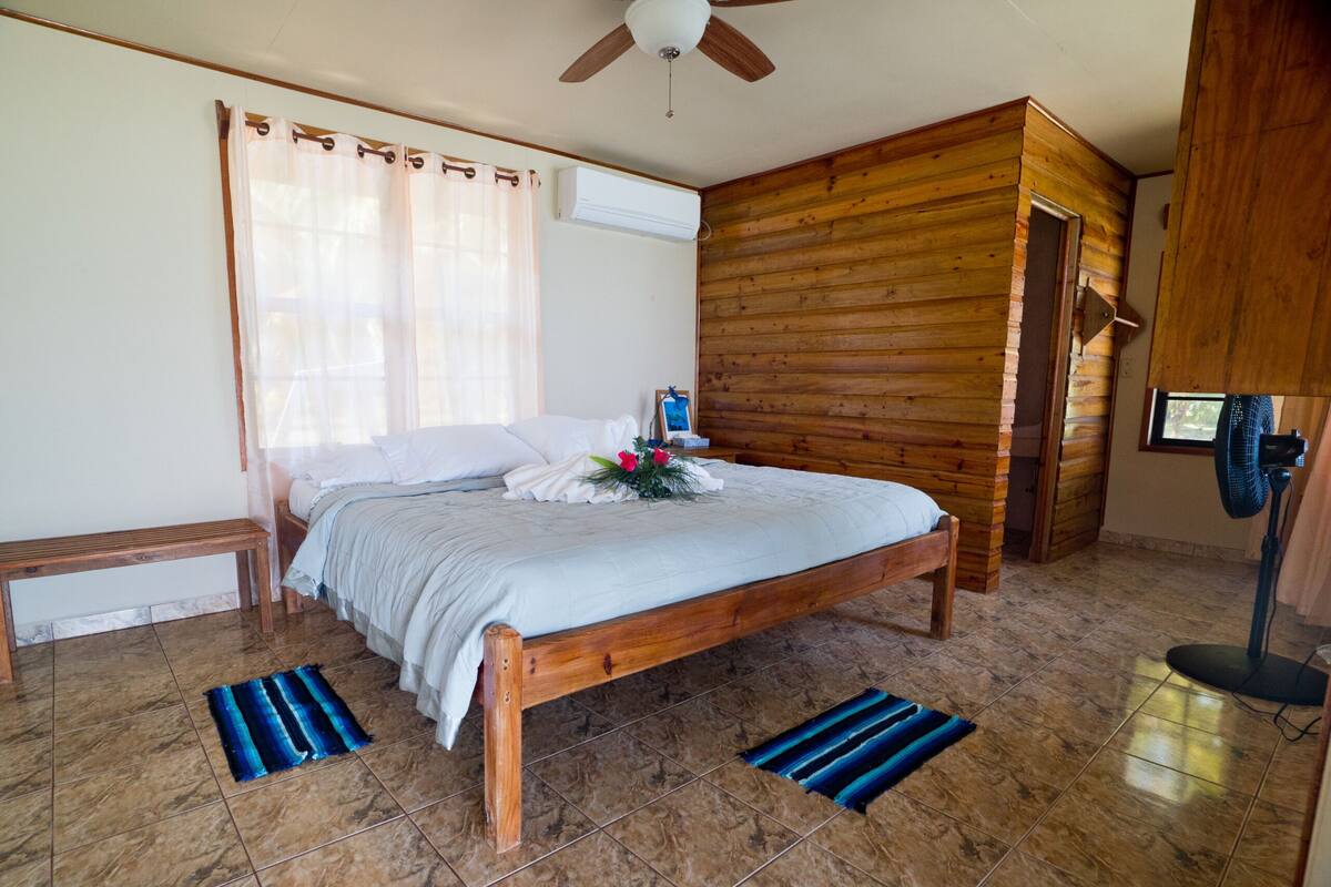 Un dormitorio con cama de madera, un banco, un ventilador de techo y una ventana con cortinas.