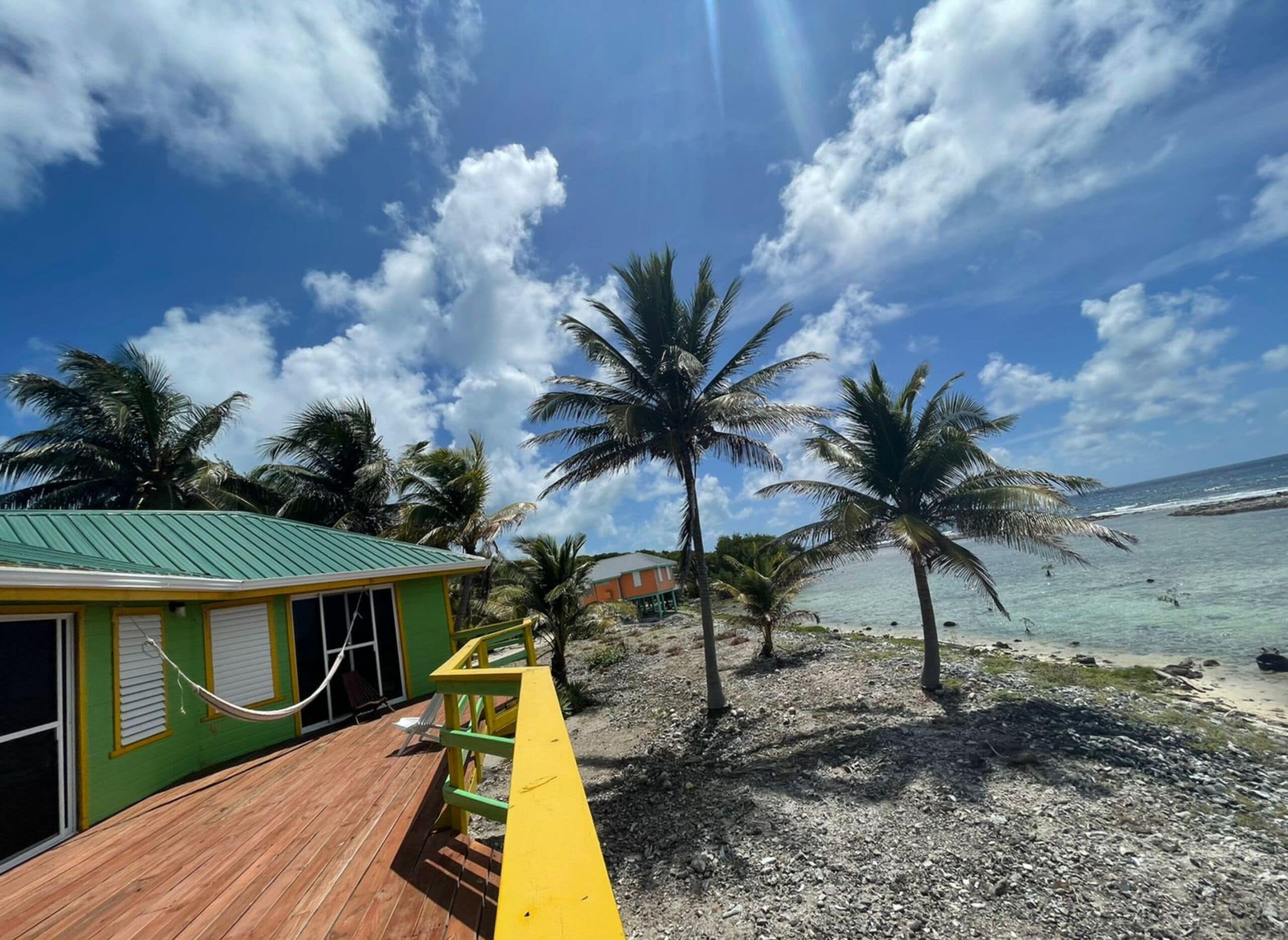 Une terrasse en bois avec une rambarde jaune, un bâtiment vert avec des volets et des palmiers au bord d’une plage.