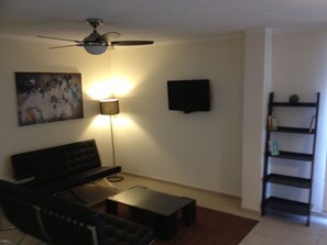 Un salon avec un ventilateur de plafond, un canapé noir, une table basse, un lampadaire, un téléviseur fixé au mur et une bibliothèque.