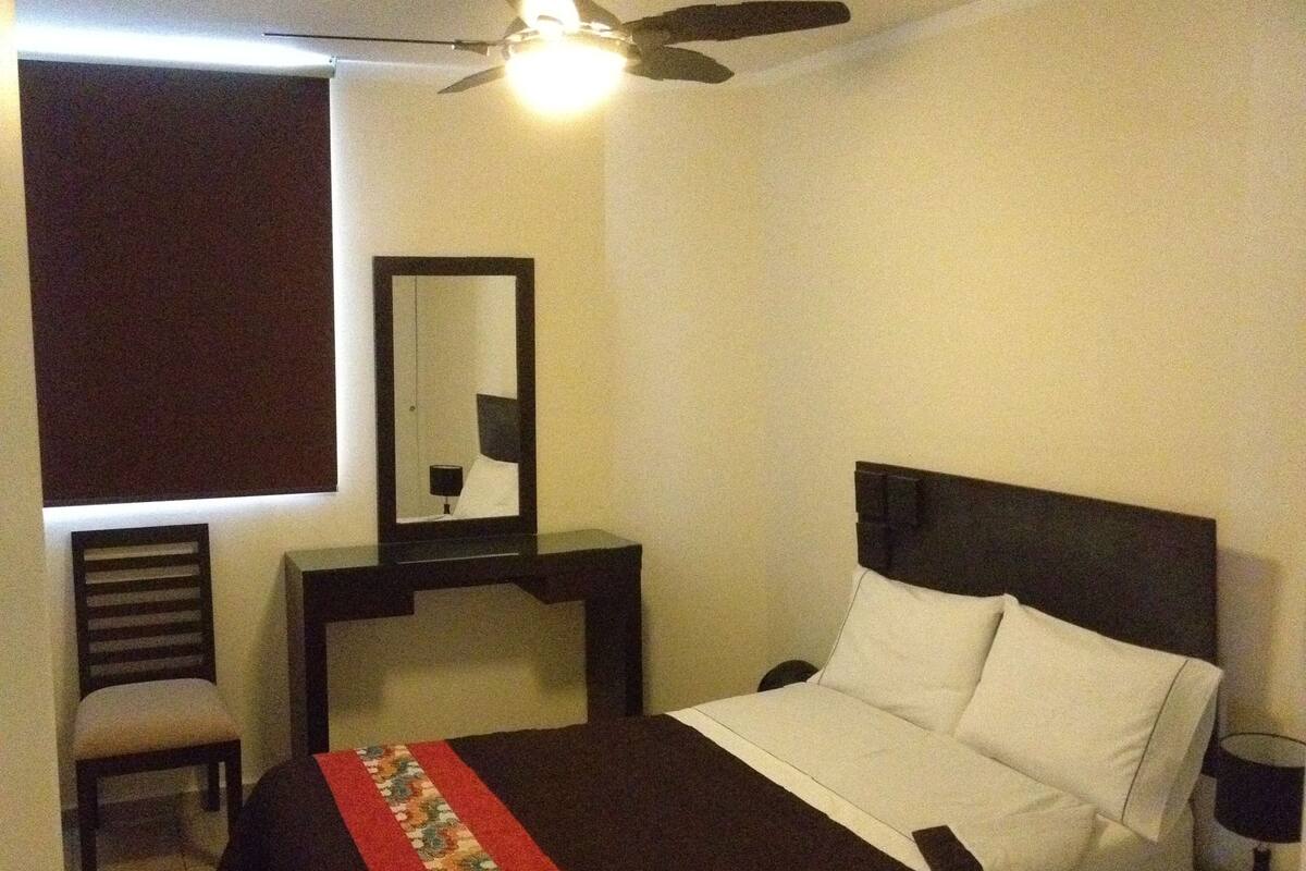 A bedroom with a bed, a chair, a nightstand, a desk, a mirror, and a ceiling fan.