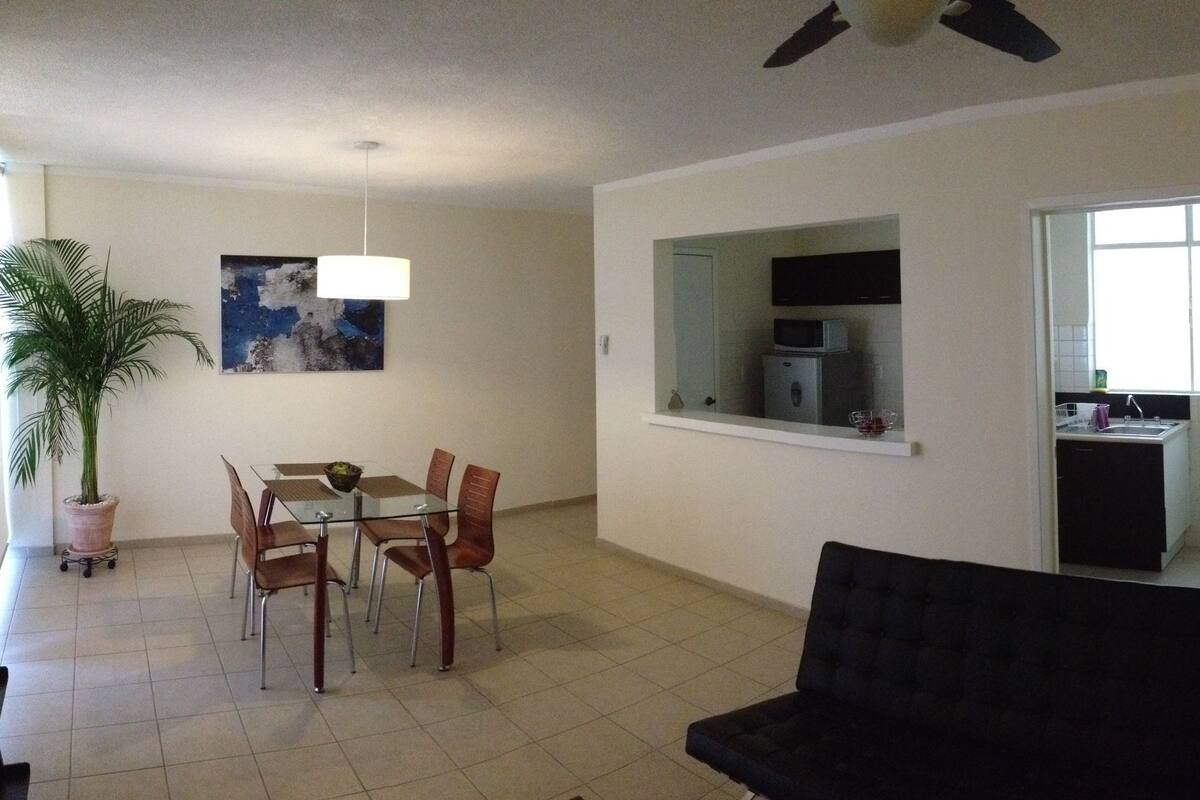A living room with a black sofa, dining table with chairs, a potted plant, and a ceiling fan.