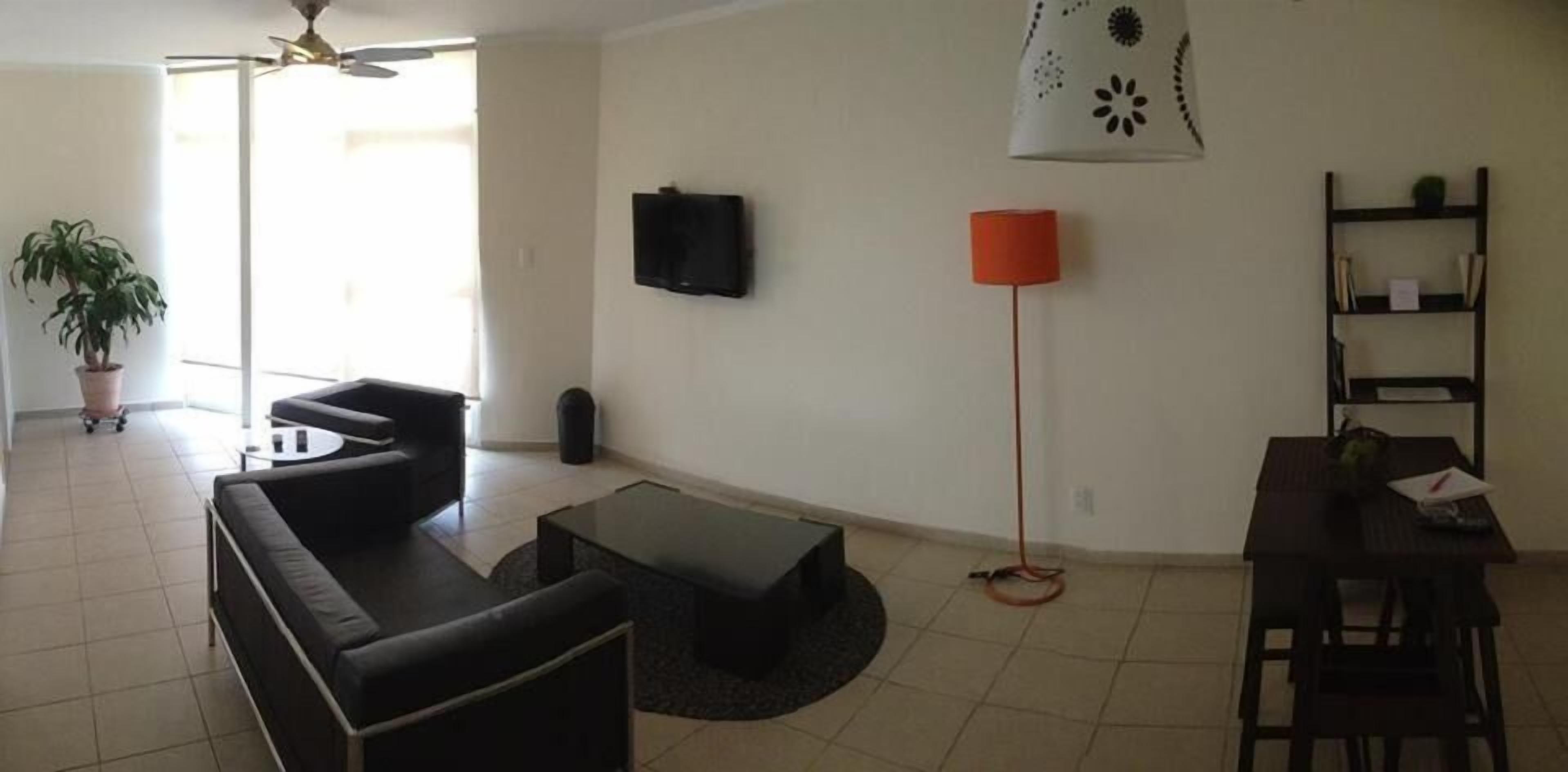 A living room with a black sofa, a coffee table, a TV mounted on the wall, a red floor lamp, and a bookshelf.