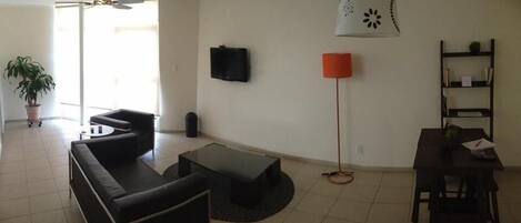 A living room with a black sofa, a coffee table, a TV mounted on the wall, a red floor lamp, and a bookshelf.