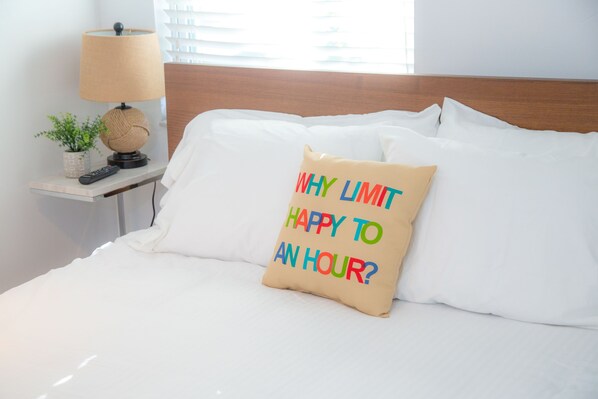 Una cama bien hecha con sábanas blancas, una lámpara, un control remoto y un cojín decorativo con texto.