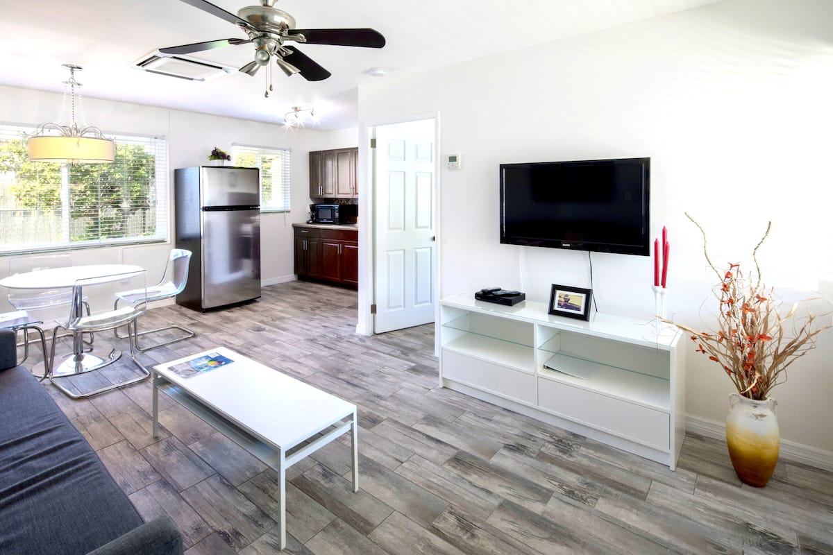 A modern living room with a white sofa, a glass coffee table, a white TV stand, and a dining area with a white table and chairs.