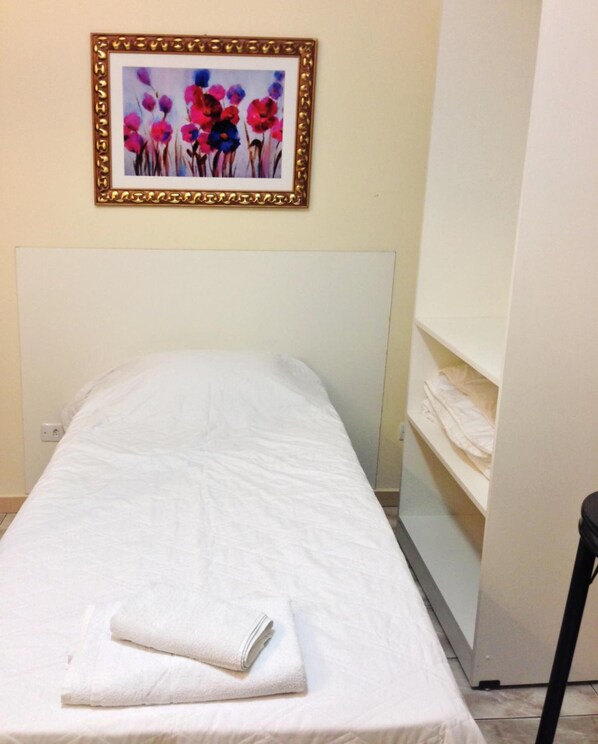 Un lit simple avec un matelas blanc et une serviette blanche pliée. Un tableau encadré représentant des fleurs est accroché au mur, au-dessus du lit.