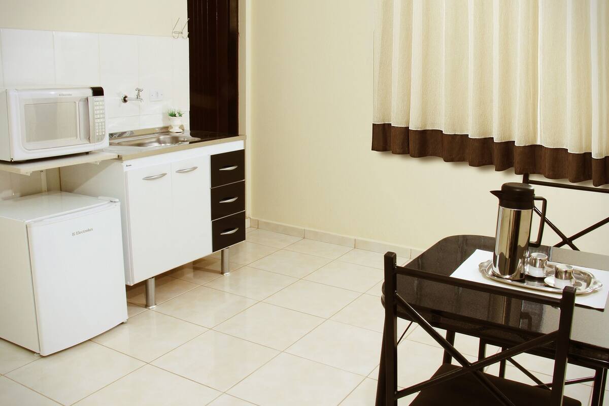 A compact kitchen with a microwave, refrigerator, and a small sink. There's a dining table with chairs and a coffee maker on the table.