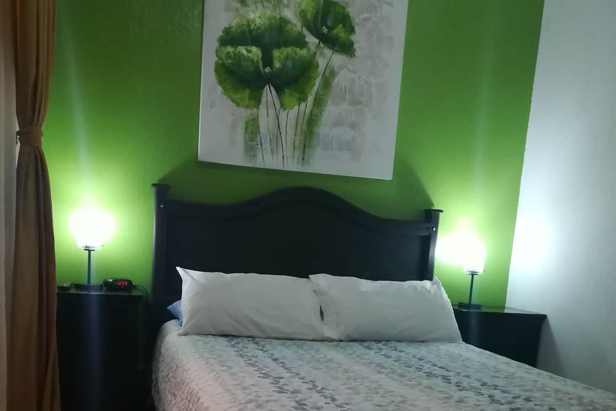 Ein ordentlich bezogenes Bett mit einem gemusterten Überwurf, zwei Nachttische mit Lampen, eine grüne Wand und ein Blumenbild.