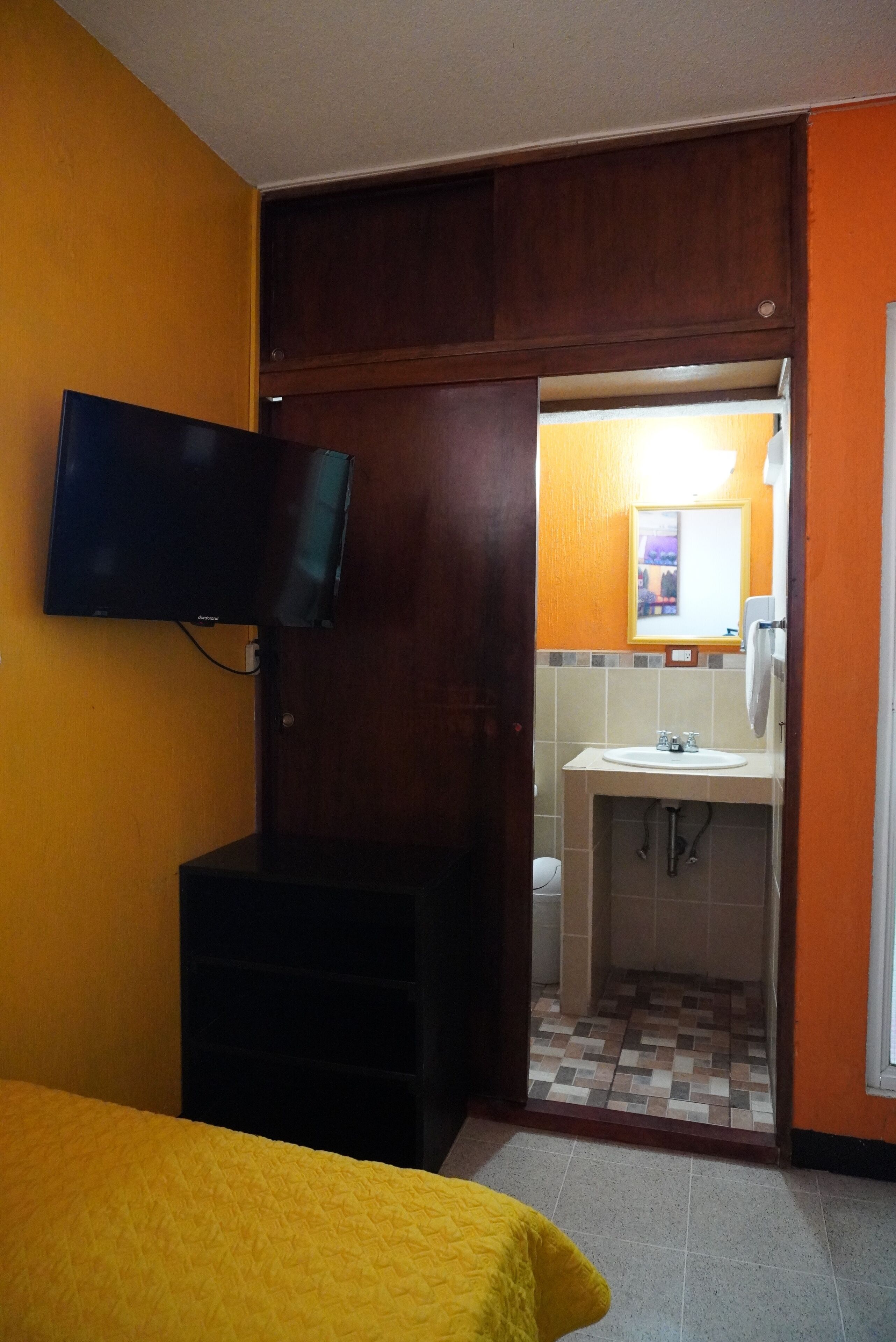 Habitación de hotel con cama, televisor montado en la pared, baño con lavabo y espejo, y suelo de baldosas.