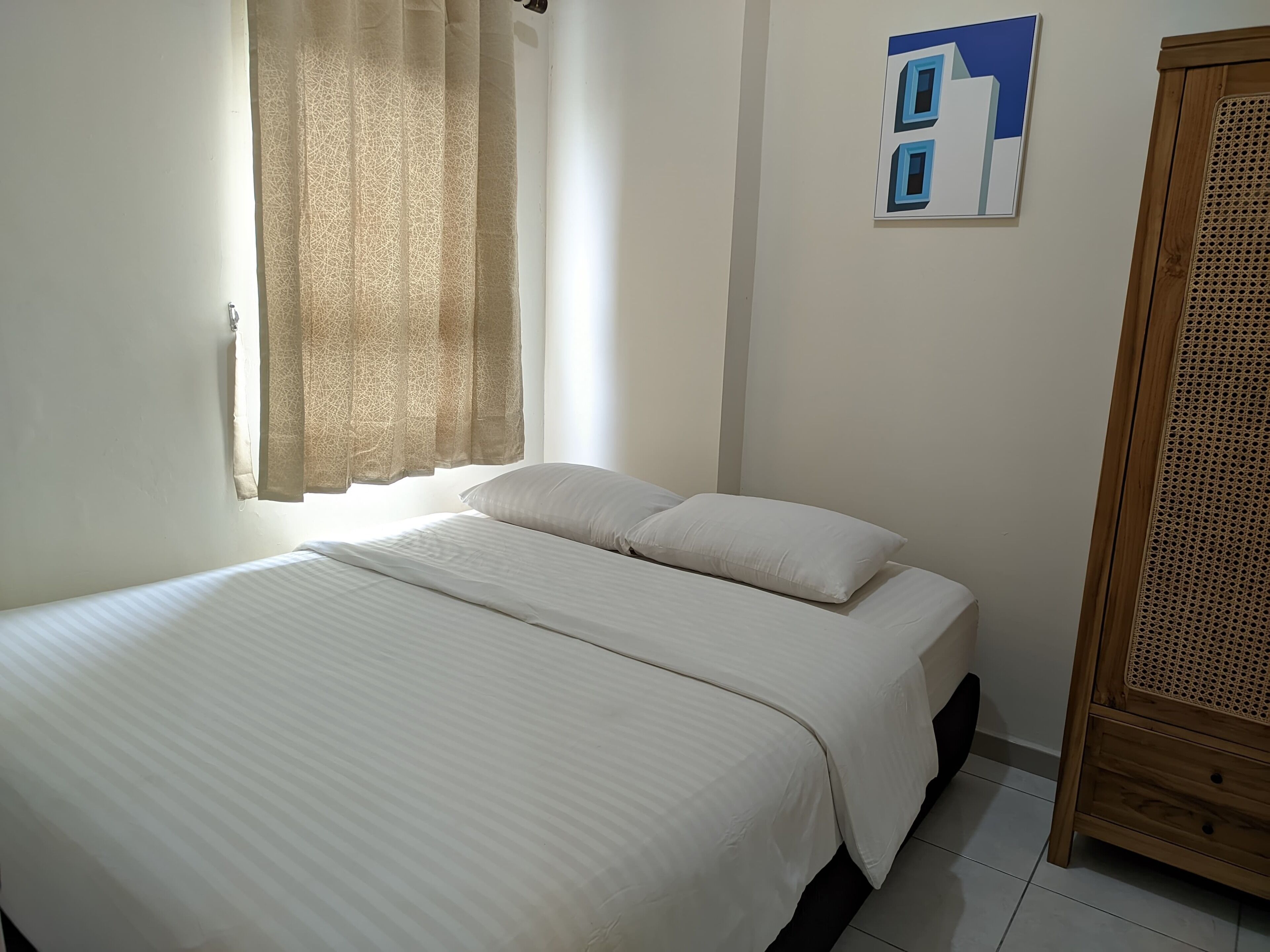 A bedroom with a bed, a window with curtains, a wall-mounted artwork, and a wooden wardrobe.