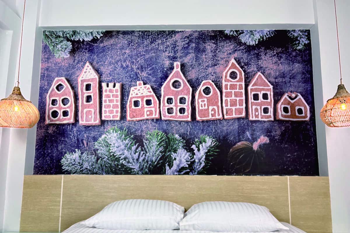 A bedroom with a bed, a wall tapestry of gingerbread houses, and two hanging lamps.