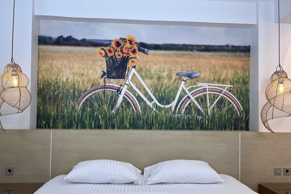 A bedroom with a bed, two white pillows, a wall-mounted painting of a bicycle in a field, and two hanging light fixtures.