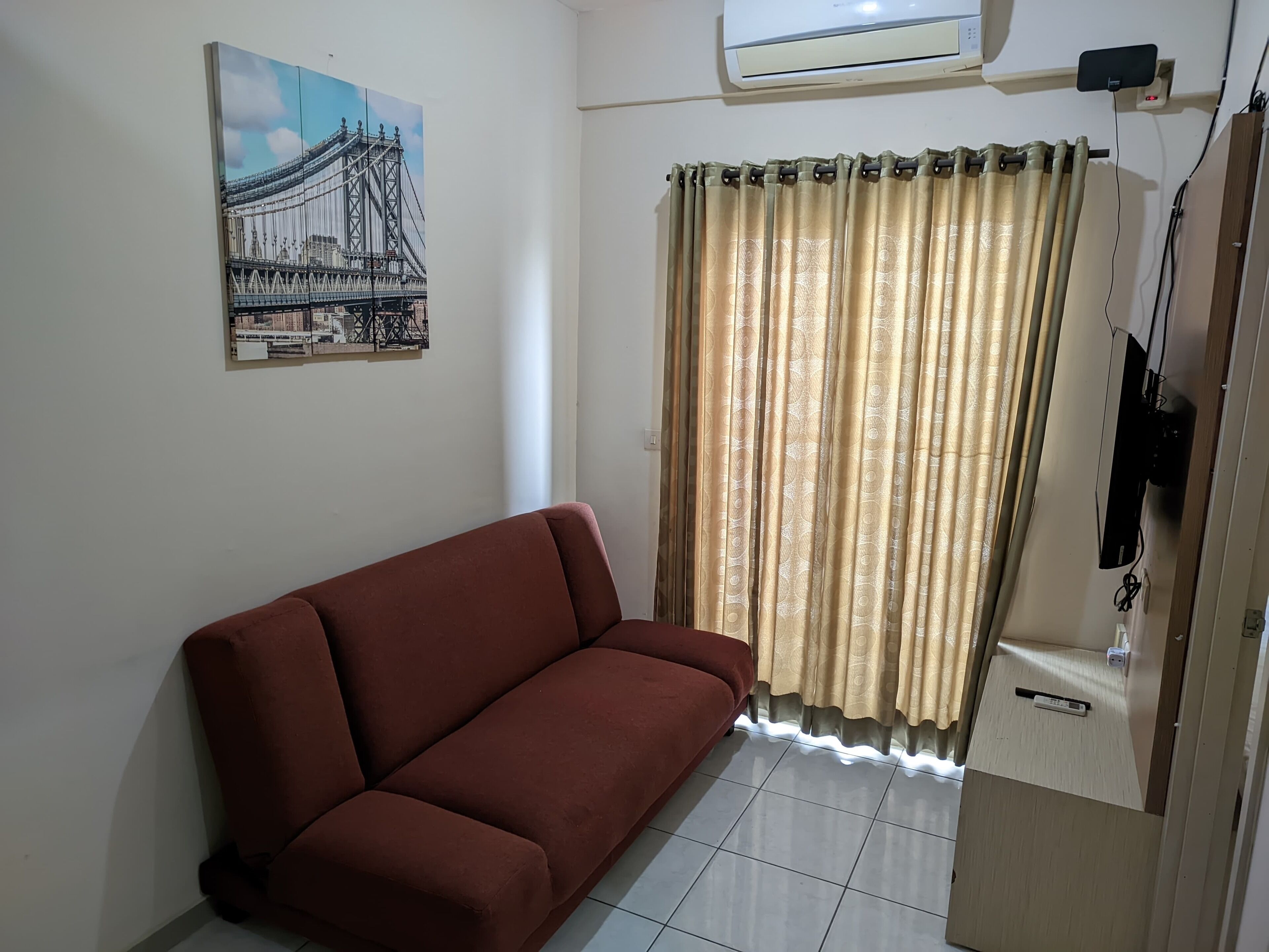 A room with a brown sofa, a large window with patterned curtains, an air conditioning unit, and a wall-mounted TV.