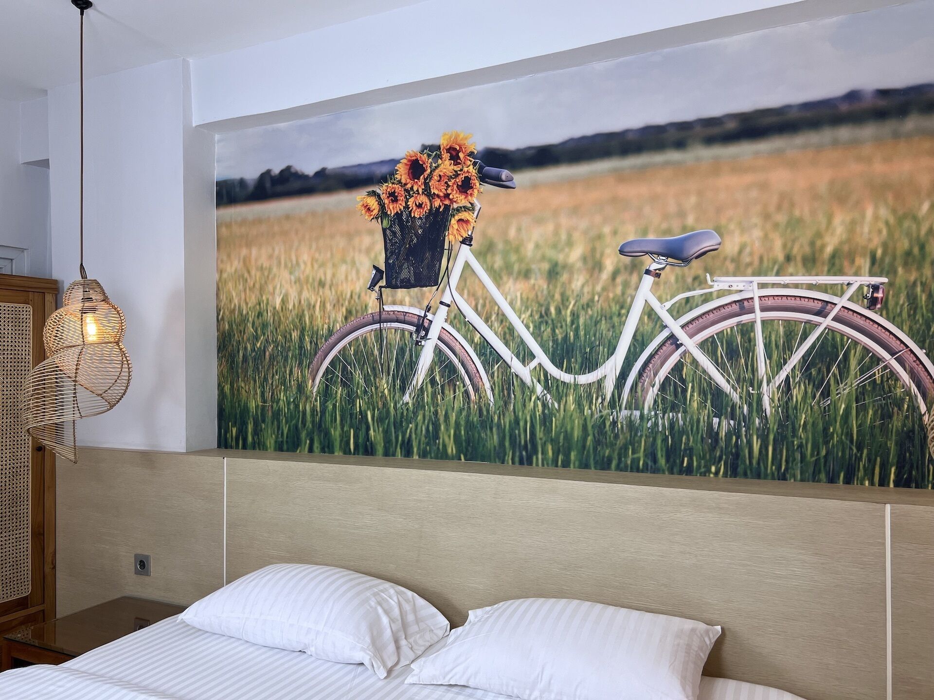 A bedroom with a bed, a wall-mounted painting of a bicycle in a field, and a wicker light fixture.