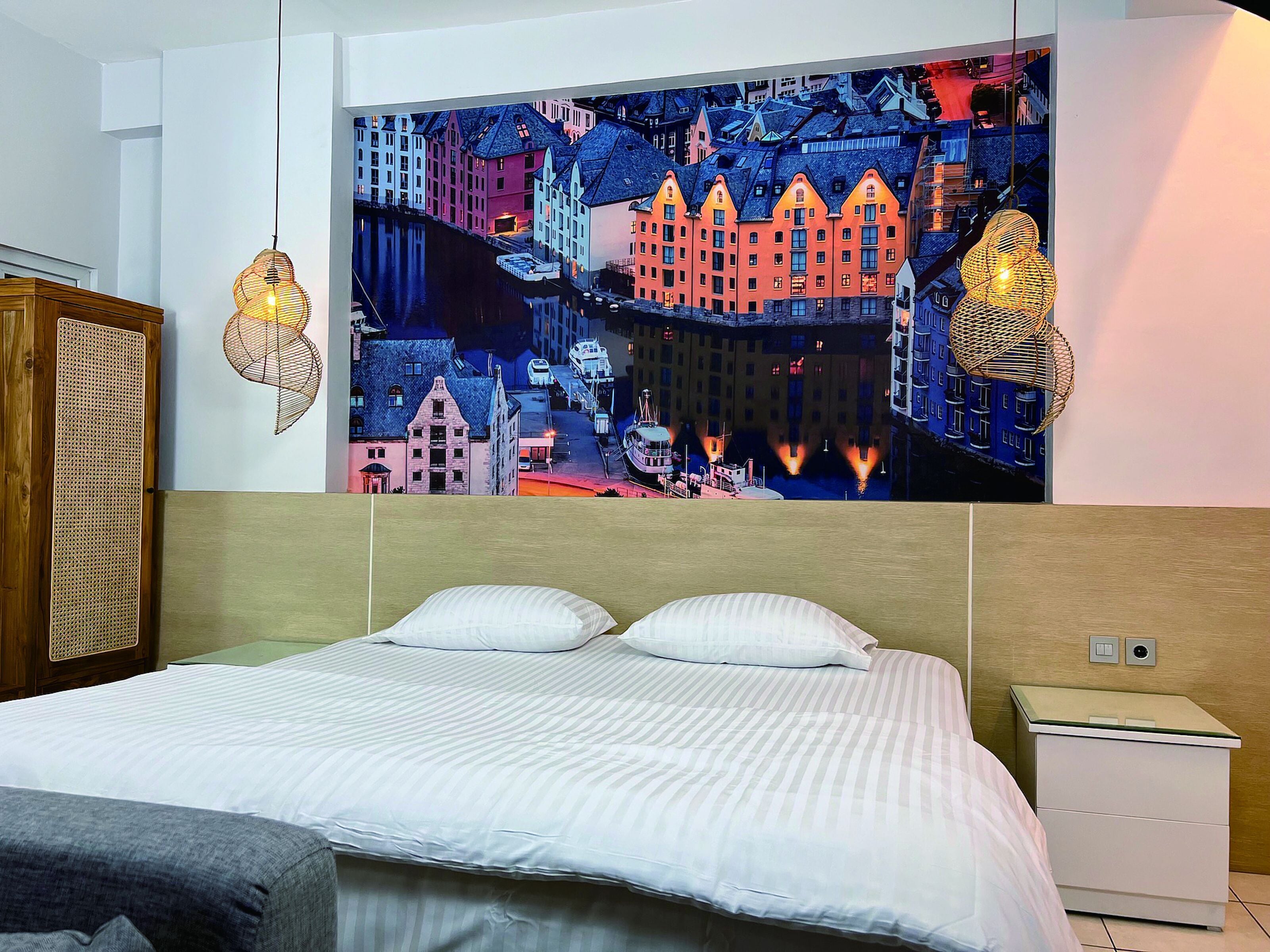A hotel room with a large bed, a nightstand, a wardrobe, and a wall mural of a cityscape.